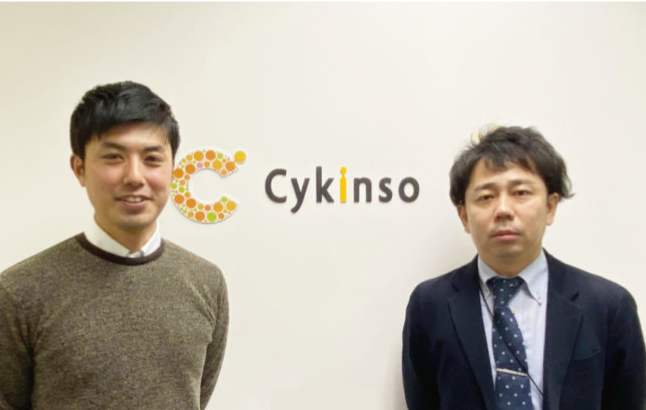 会社のロゴ前で二人の男性が並んでいる様子