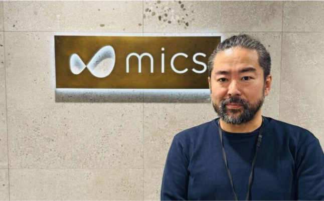 会社のロゴ前で髭を生やした男性が立っている様子
